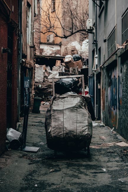 A narrow urban alleyway filled with overflowing rubbish and discarded materials, with a large black garbage bag in the foreground, placed on the dirty, uneven pavement. Behind the bag, there are stacks of cardboard boxes, plastic bags, and miscellaneous trash piled up against a cluttered background of graffiti-covered walls and exposed brick facades. To the right, the side of a building features metal window grilles and small air conditioning units, while the left side shows a weathered door and additional debris scattered across the ground. Bare tree branches are visible at the top, suggesting an outdoor setting in a city environment. The scene is dimly lit, with natural light filtering through, creating shadows that highlight the mess and density of waste typical of a site requiring waste collection or alternative rubbish removal services, such as those offered by Rubbish Removal Shoreditch, which manages on-site clearance for residents and traders alike.