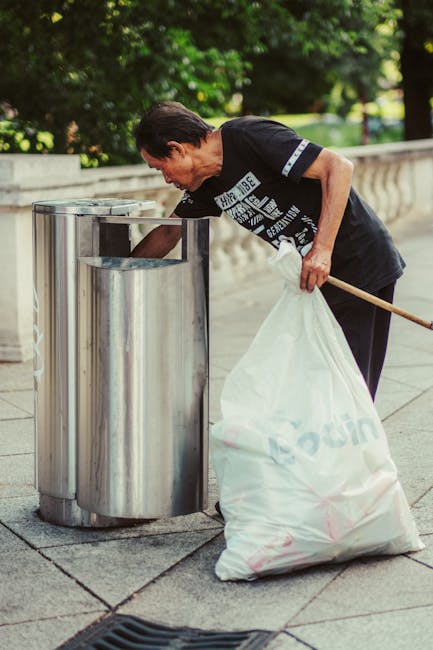 A middle-aged man with dark hair, wearing a black T-shirt with white text and graphics, is seen standing on a paved sidewalk during daytime, leaning forward to empty a silver, cylindrical metal trash bin with an open top. In his left hand, he holds a large, partially transparent white plastic bag filled with rubbish, and his right hand is reaching into the trash bin. The scene is set outdoors with lush green trees and foliage in the background, and a short stone wall runs parallel to the sidewalk. The environment appears clean and well-maintained, suggesting a routine outdoor waste management activity, possibly part of private rubbish collection or on-site clearance, carried out by Rubbish Removal Shoreditch. The overall atmosphere is neutral and professional, emphasizing practical rubbish disposal methods suited for flats or estates where independent waste handling might be preferred over local authority collection.