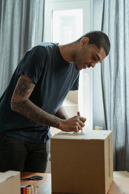 A young man with short dark hair and medium skin tone is focused on writing or marking on a cardboard box using a red pen. He is dressed in a dark grey t-shirt and is standing in a well-lit room near a large window with light blue or grey curtains, which diffuse natural daylight. The surface of the cardboard box appears to be a standard brown corrugated material. The environment suggests an indoor setting, possibly a home or workshop, where individual or private waste handling might take place before disposal or collection. The scene emphasizes careful handling of packaging, which is relevant to alternatives to regular rubbish collection, such as on-site clearance or private waste disposal services, aligning with the context of rubbish removal and waste management support provided by Rubbish Removal Shoreditch.