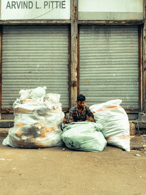 A man sitting on the ground between two large, transparent plastic bags filled with mixed waste materials, including dark-colored and light-colored items, positioned in front of a closed, weathered metal roller shutter with rusted vertical supports. The man, with dark hair and a beard, wears a patterned shirt and appears to be resting or waiting, surrounded by additional smaller green and white rubbish bags. The scene takes place on a paved street surface, with a worn wall above the shutter featuring a partially visible sign. The overall environment suggests an area where private waste collection or on-site rubbish disposal activities are taking place, associated with independent rubbish removal services such as those offered by Rubbish Removal Shoreditch.